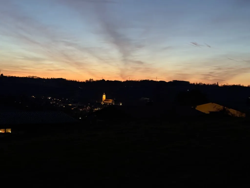 Blick über ein dunkles Tal auf einen beleuchteten Kirchturm in der Abenddämmerung