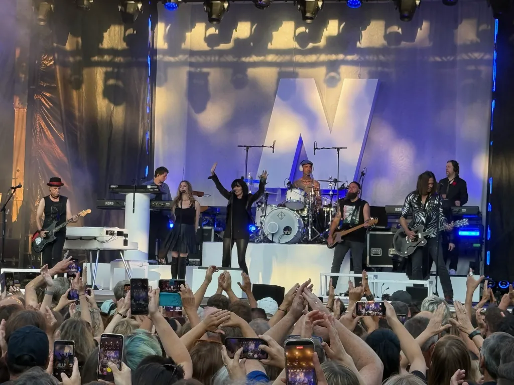 Nena mit erhobenen Armen auf einer Konzertbühne mit Band, davor ein Publikum mit Handys in der Luft.