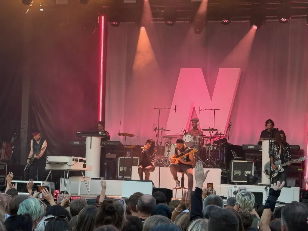 Band auf einer pink beleuchteten Bühne mit großem „N“ im Hintergrund, davor ein Publikum mit erhobenen Händen.