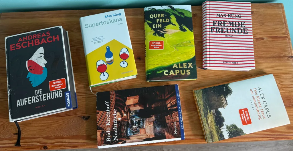 Sieben Bücher auf einem Holztisch, darunter „Die Auferstehung“, „Supertoskana“, „Querfeldein“ und „Fremde Freunde“.