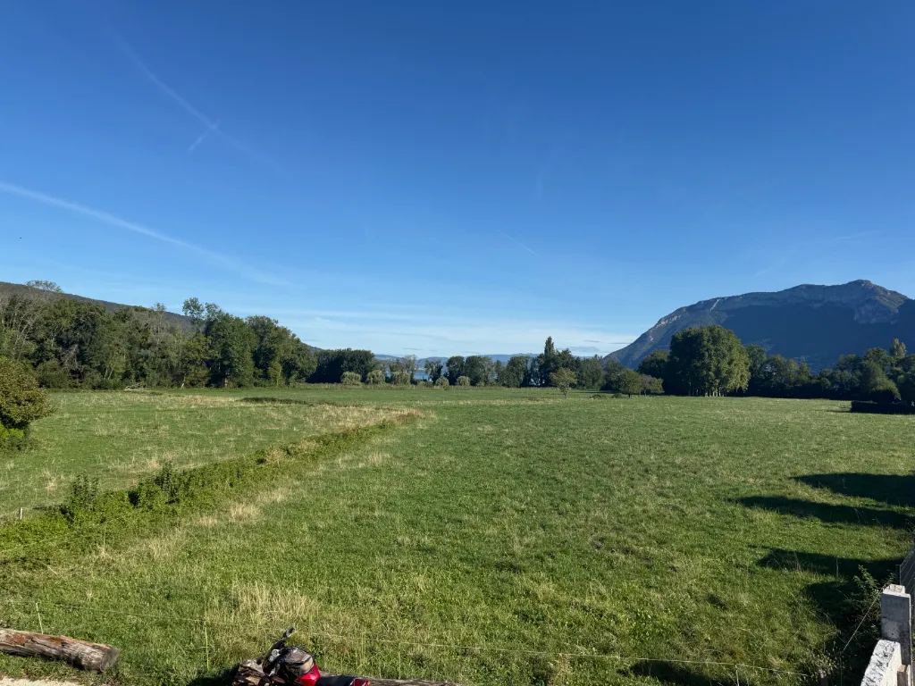 Weite grüne Wiese mit Baumreihe, dahinter ein See und Berge unter blauem Himmel, vorn ein Motorrad am Zaun.