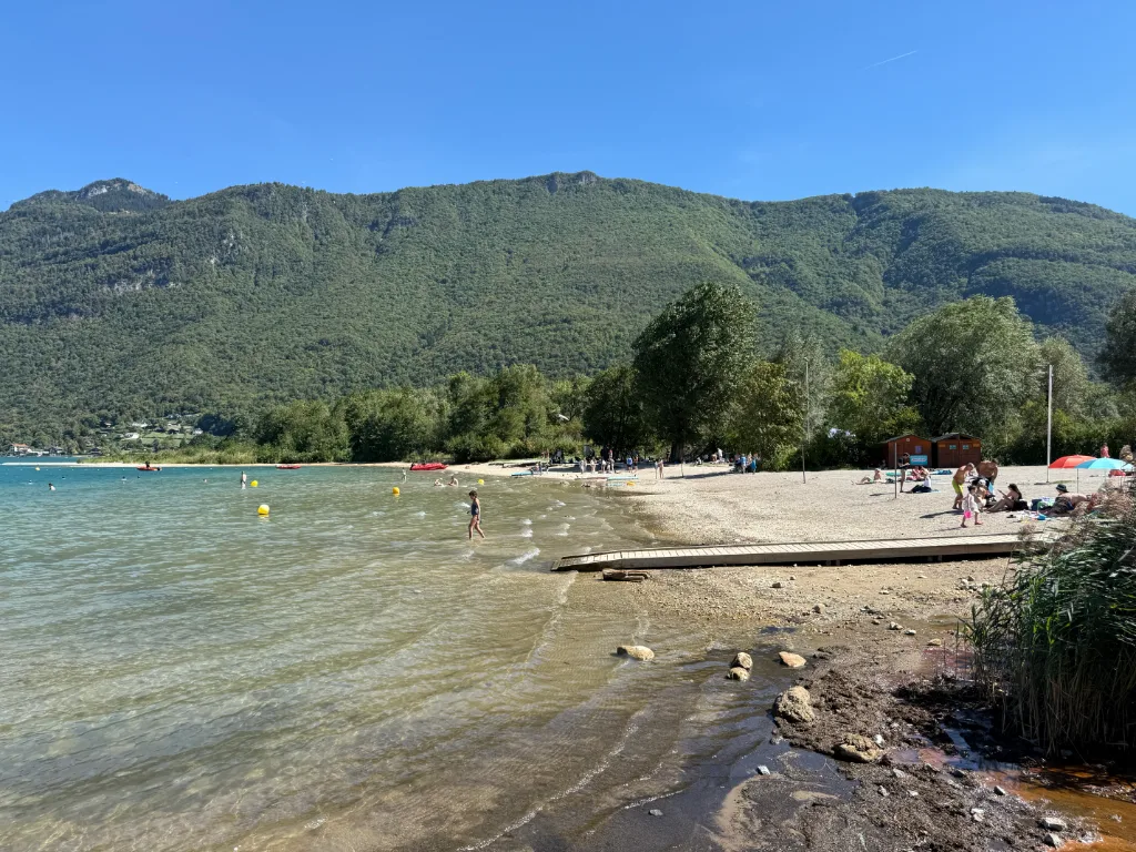 Badegäste an einem flachen Kiesstrand mit Holzsteg an einem klaren Bergsee vor bewaldeten Berghängen.