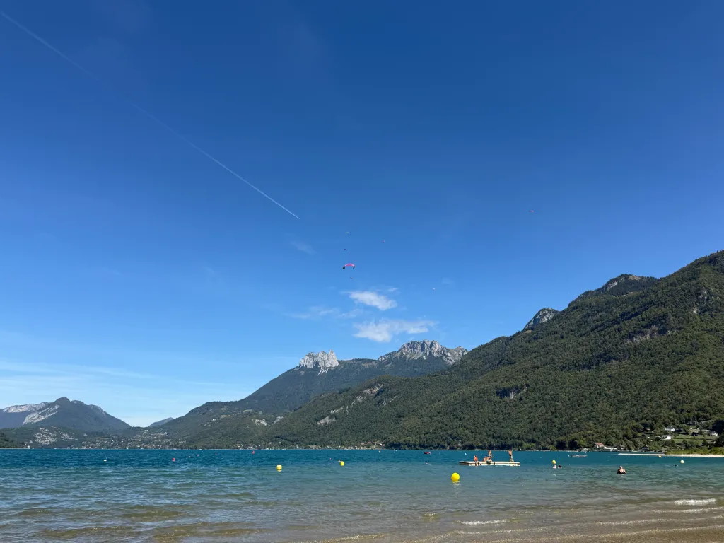Türkisfarbener Bergsee mit Badeplattform, gelben Bojen und Gleitschirmen über bewaldeten Berghängen bei blauem Himmel.