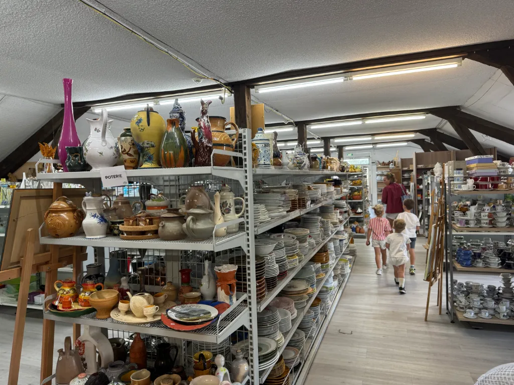Volle Regale mit Geschirr und Keramik in einem Gebrauchtwarenladen, Schild „Poterie“, im Gang Kinder und eine Frau.