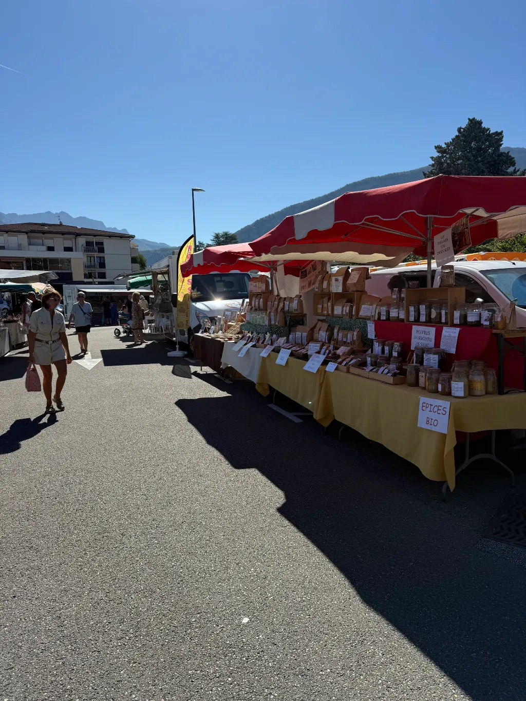 Marktstand mit Gewürzen unter rotem Sonnenschirm, Schild „EPICES BIO“, links geht eine Frau vorbei.