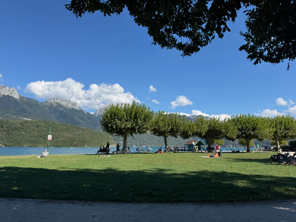 Menschen entspannen auf einer Liegewiese am türkisblauen See, dahinter eine Baumreihe und Berge unter blauem Himmel.