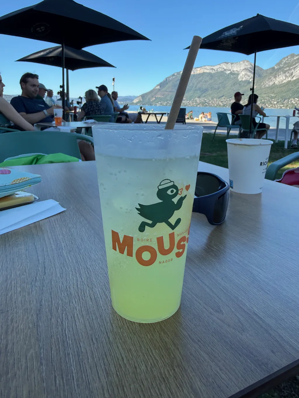 Gelbes Getränk mit Papierstrohhalm im Becher mit Enten-Logo auf Tisch einer Strandbar am See vor Bergen.
