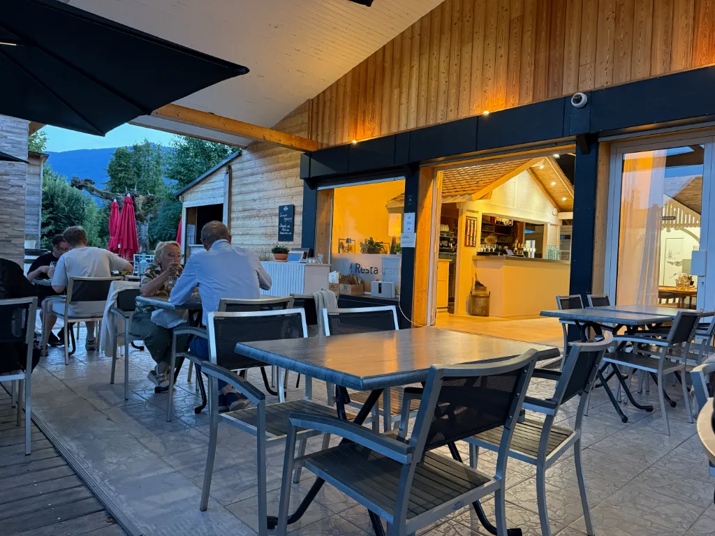 Überdachte Restaurantterrasse in der Dämmerung, links sitzen Gäste an Tischen, rechts der beleuchtete Innenraum.
