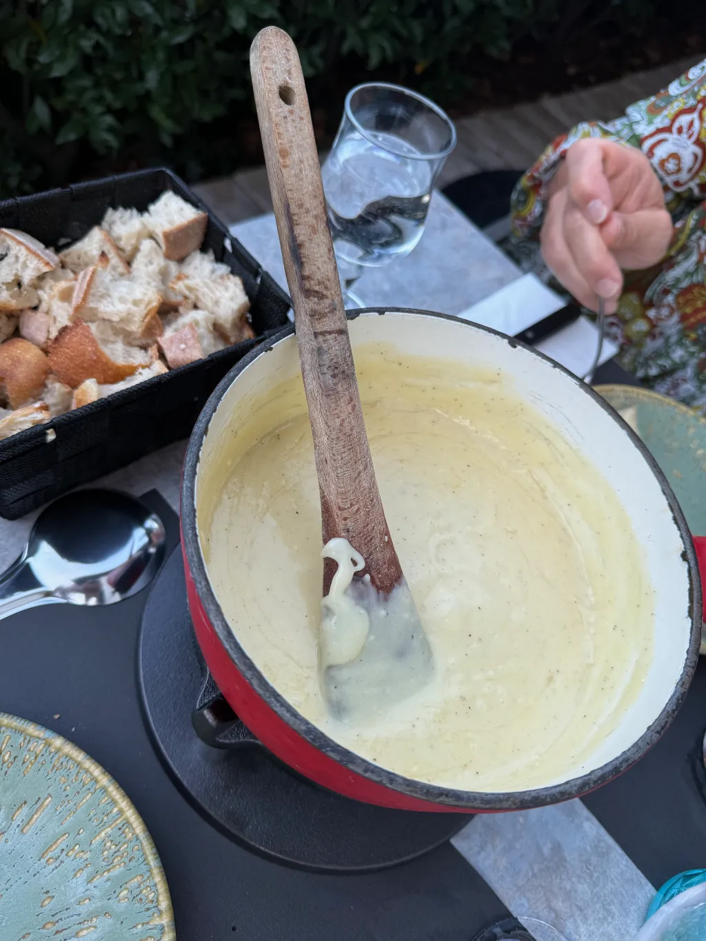 Roter Topf mit geschmolzenem Käsefondue und Holzkelle, daneben ein Korb mit Brotwürfeln auf dem Tisch.
