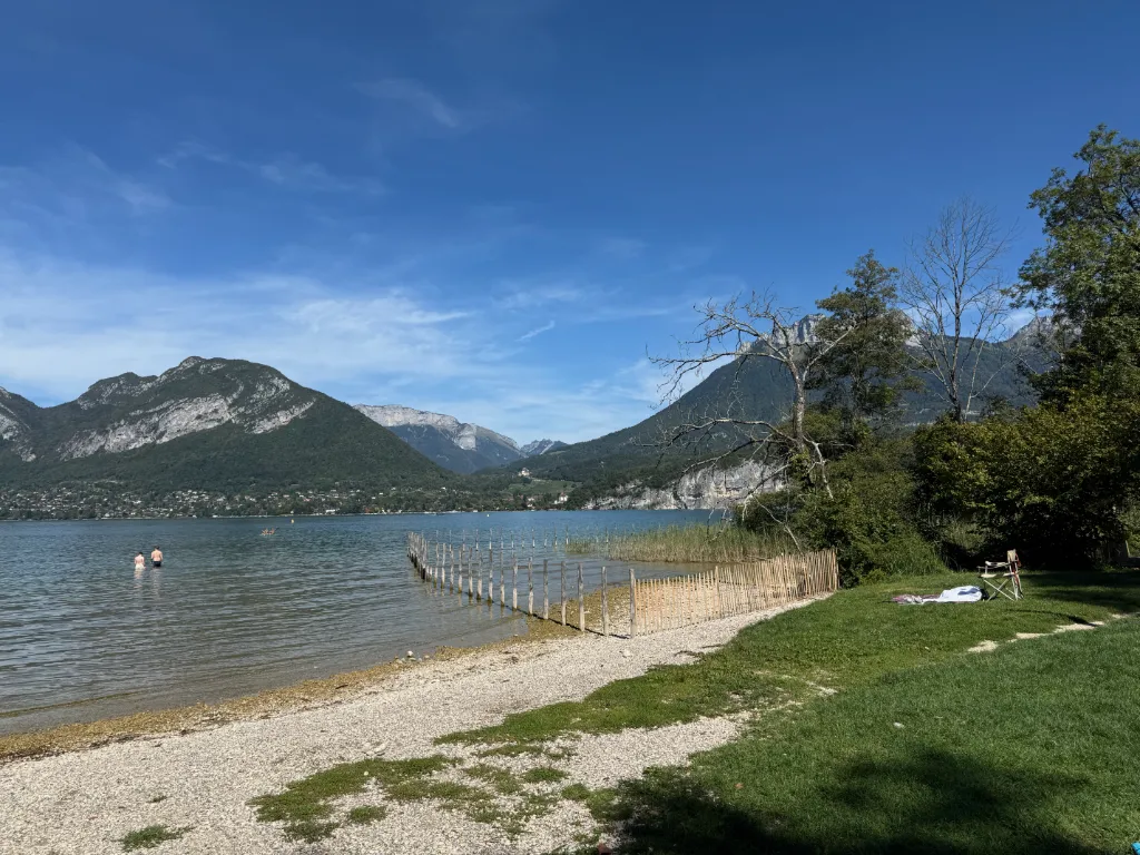Zwei Personen stehen im flachen Wasser eines Bergsees mit Kiesstrand, Holzzaun und bewaldeten Gipfeln.