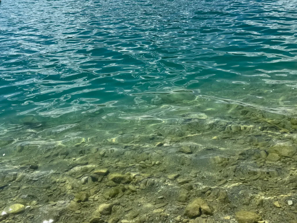 Klares türkisgrünes Wasser mit Steinen im flachen Uferbereich, das nach hinten dunkler und tiefer wird.