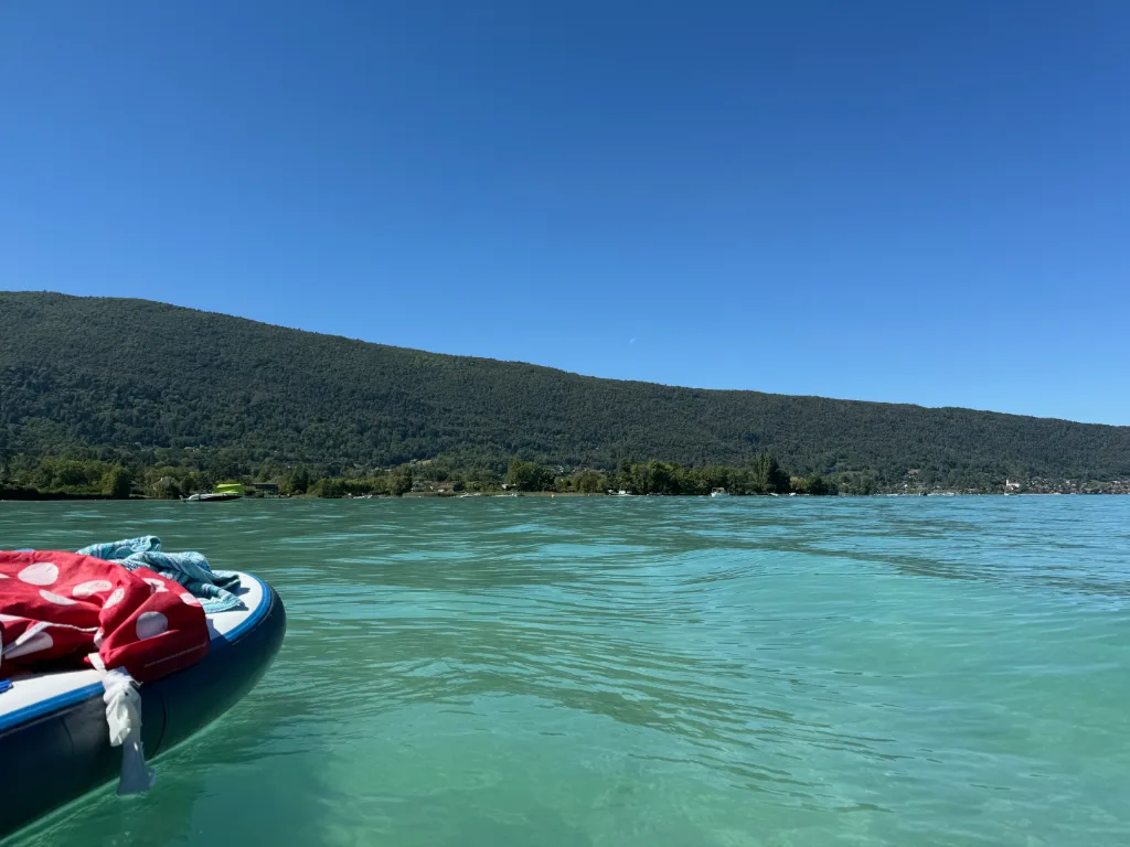 Blick von einem Stand-up-Paddleboard mit Handtüchern über türkisgrünes Wasser auf bewaldete Hügel unter blauem Himmel.