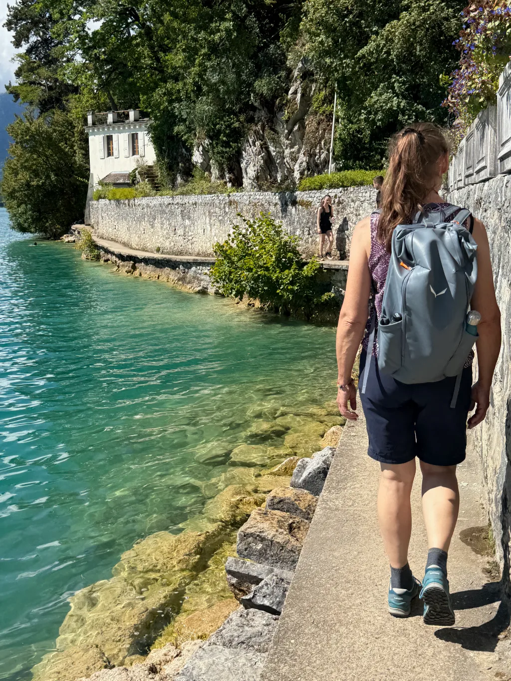 Frau mit blauem Rucksack geht auf schmalem Uferweg entlang eines türkisfarbenen Sees mit Mauer und Bäumen.