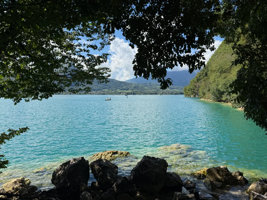 Blick durch Laubzweige auf einen türkisfarbenen Bergsee mit Felsen am Ufer und bewaldeten Hügeln.