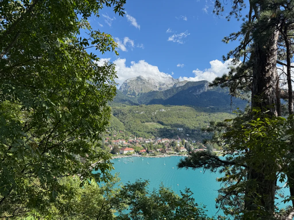 Blick durch Bäume auf einen türkisfarbenen Bergsee mit Segelbooten, Uferort und bewaldetem Gebirge.